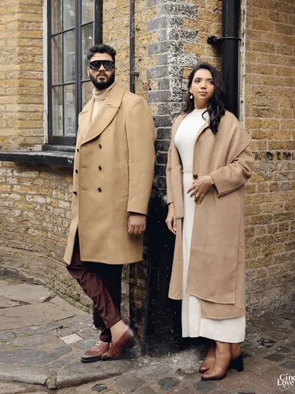 Posing against a rustic brick wall in London. The contrast between his burgundy trousers and their beige coats adds a subtle pop of color to the otherwise neutral palette.