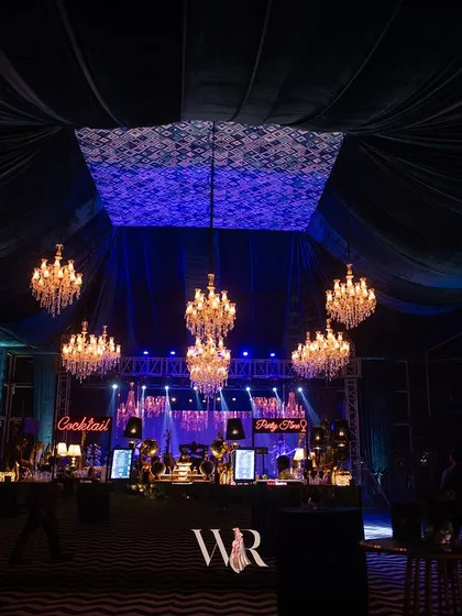 A wide shot of the bar and stage area for a modern cocktail night. The design uses cool blue lighting, a custom ceiling installation, and a striking chevron floor to create a high-energy, club-like atmosphere.