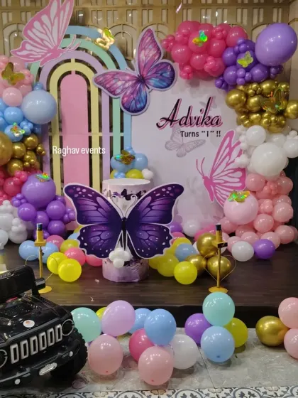 A butterfly themed first birthday party with a toy jeep for photos. The setup includes a colorful backdrop, balloon decorations, and a large light-up number '1'.