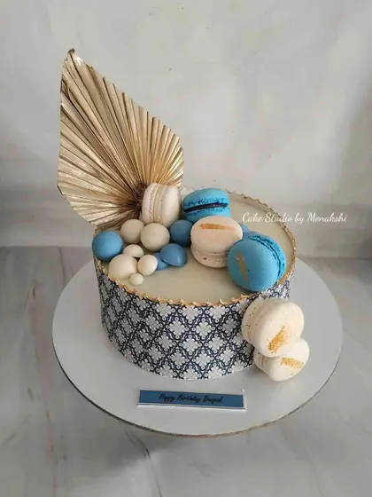 A stylish no-fondant cake in a blue and white theme. The design features a printed edible sheet, macarons, and a dried palm spear for a modern and chic look.