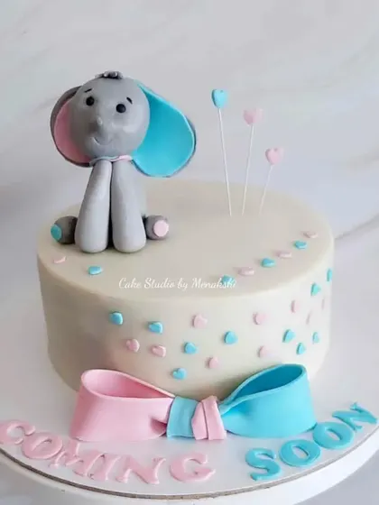 A cute baby shower cake with a "Coming Soon" message. The design features an adorable elephant and a pink and blue bow, perfect for a gender reveal.