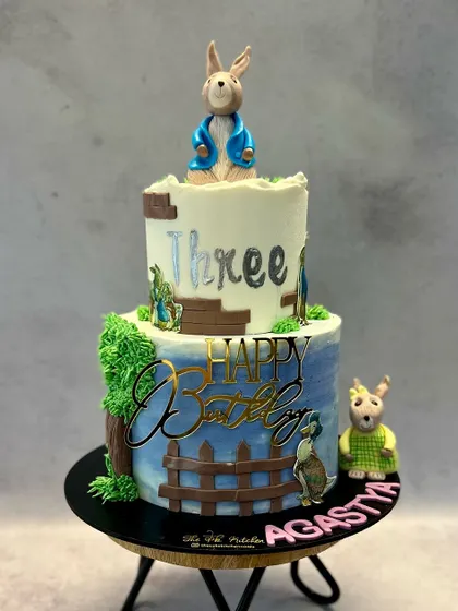 A charming two-tiered Peter Rabbit cake, perfect for a classic storybook-themed party. The hand-painted watercolor background and fondant figures bring the garden scene to life.