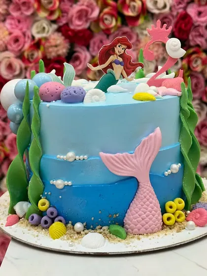 A vibrant mermaid cake with a sculpted Ariel topper, a pink flamingo float, and colorful underwater elements. It's a playful and imaginative design.