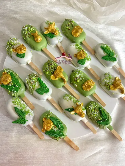 A close-up of my jungle-themed cakesicles. I use a mix of green and white chocolate coatings, detailed with edible gold animal figures, delicate leaf patterns, and sprinkles to make each one a tiny work of art.