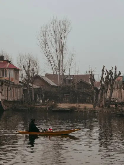 A solitary boatman against a backdrop of traditional Kashmiri houses and bare Chinar trees, creating a moody, wintery scene.