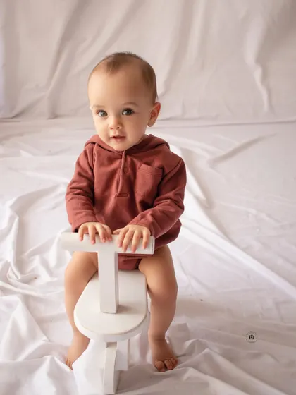 A simple and classic sitter portrait. A baby in a rust-colored romper sits on a small white toy bike against a clean backdrop.