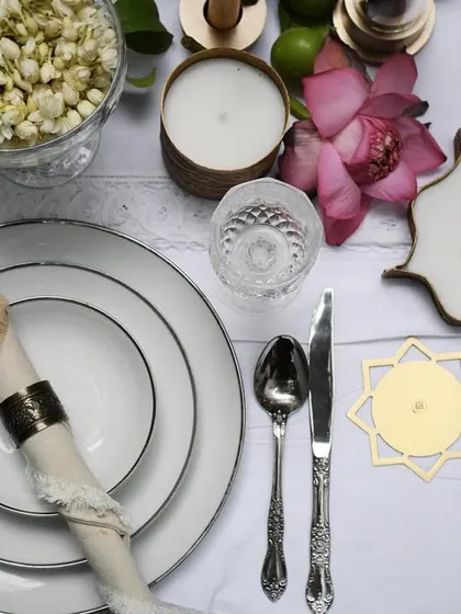 An overhead flat lay of a festive table setting for Byaas Home. This detailed shot highlights the beautiful interplay between the crisp white dinnerware, intricate silverware, and the brand's unique star-shaped candles and coasters.