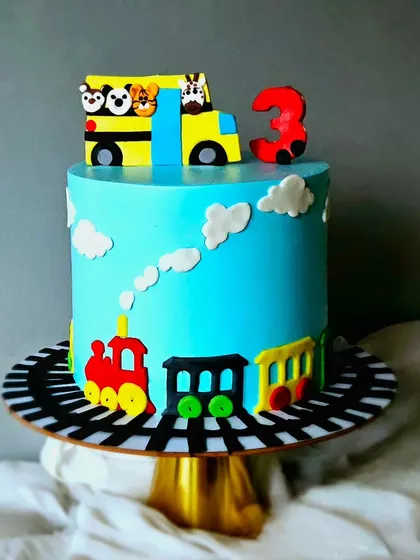 A cute bus and train theme cake for a third birthday. This design features a bright blue whipped cream cake with a fondant train running along the base and a school bus topper.