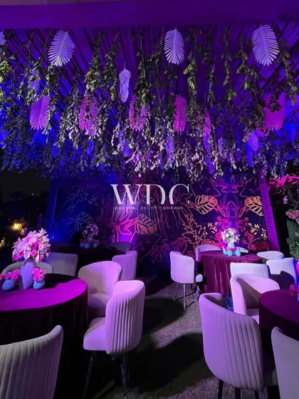 A chic lounge area for the birthday party. The space features a ceiling of hanging greenery and leaves, with comfortable seating and a moody, purple-lit ambiance.