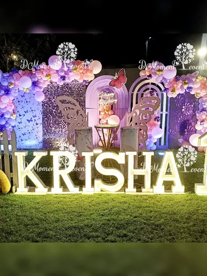 An enchanting outdoor evening setup for a butterfly-themed first birthday, featuring a sequin wall, balloon arches, and illuminated letters on a lush green lawn.
