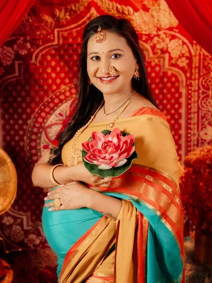 A smiling portrait of a mom-to-be in a colorful saree, holding a lotus flower. The rich red background and traditional jewelry complete this vibrant and happy maternity photo.