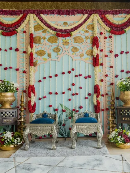 A beautiful setup for an intimate engagement, featuring ornate silver chairs against a backdrop of hanging floral strings and a custom-painted panel. The decor is elevated by traditional brass pots and floral arrangements.