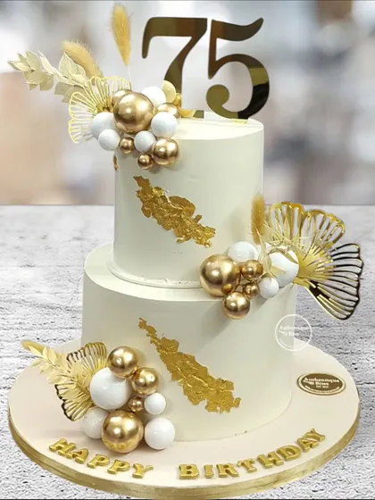 A stylish two-tiered cake celebrating a 75th birthday. The design uses white and gold spheres and dried palm leaves for a modern and sophisticated milestone cake.