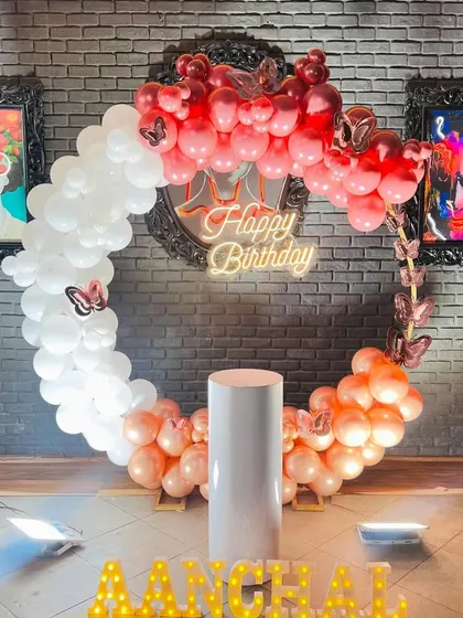 A stylish circular balloon arch in red, white, and rose gold, perfect for a modern birthday party. The arch is accented with butterfly details and a central neon sign.