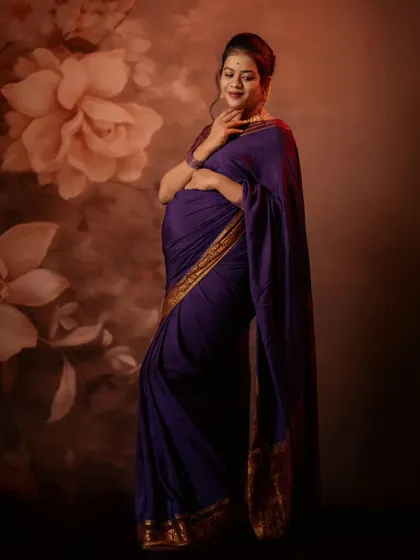 Draped in a stunning purple saree, this mother-to-be poses elegantly against a floral studio backdrop, showcasing a timeless and graceful maternity look.