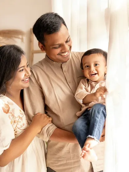 A family of three enjoying the light from a large window. The baby's happy expression and the parents' smiles create a warm and loving scene.