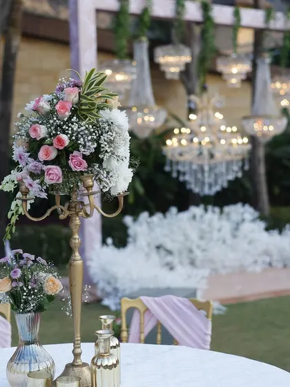 An elegant table setup for an outdoor wedding. A floral centerpiece with soft pink and white roses sits on the table, with grand crystal chandeliers hanging in the background, blending garden romance with pure luxury.