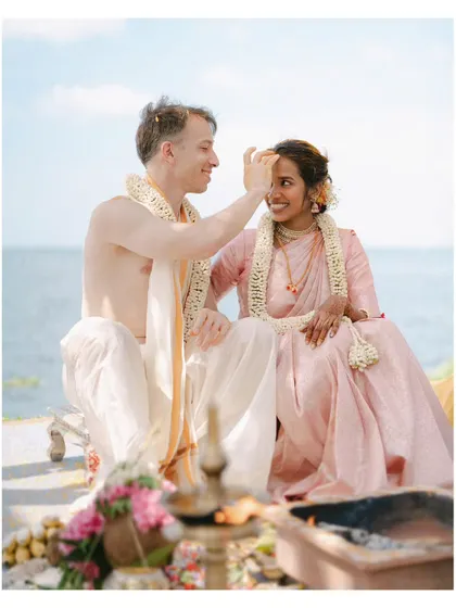 A tender moment during a seaside Hindu ceremony, as the groom applies a tilak to the bride's forehead. The sea behind them adds a sense of infinite possibility to their union.