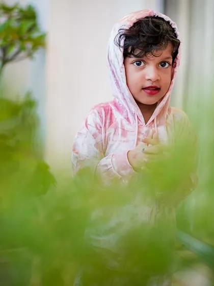 A beautiful portrait of a young girl in a pink velour hoodie, captured through the foliage of balcony plants, adding depth and a sense of wonder.