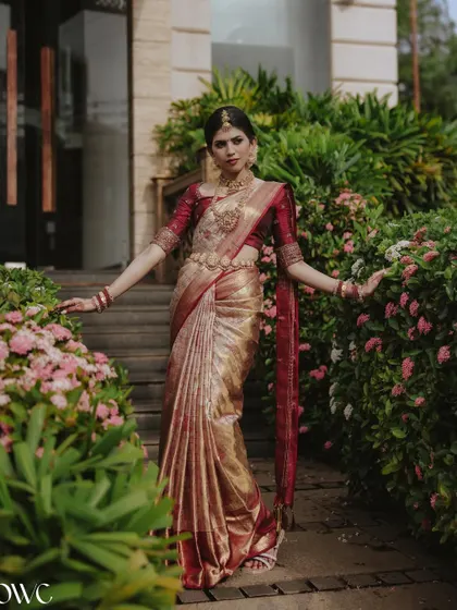 A full-length portrait of the bride amidst lush greenery and flowers, her red and gold Kanjeevaram saree looking absolutely radiant.