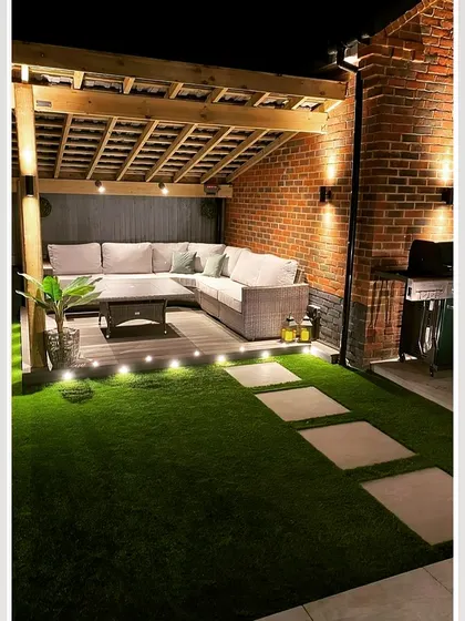 A cozy backyard corner featuring a wooden pergola attached to a brick wall. The space is transformed into an intimate lounge with a large sectional sofa and ambient lighting from string lights and ground-level spotlights.