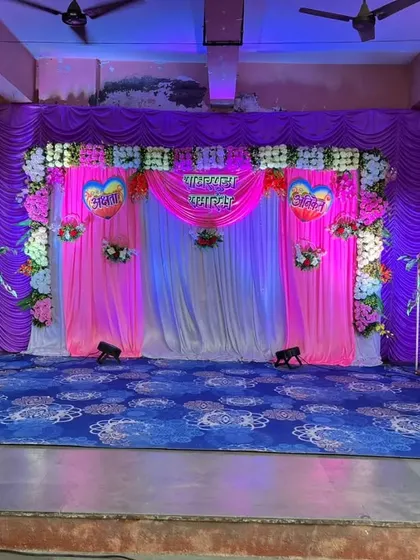 A colorful stage setup in a community hall, using bright pink and purple drapes. The simple floral arrangements and heart-shaped cutouts make it a budget-friendly yet festive option for engagements or family functions.