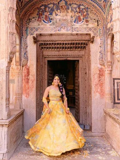 The bride twirls joyfully in her yellow lehenga in front of an ancient, ornate doorway, her happiness lighting up the historic space.