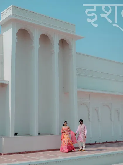 A minimalist and regal shot of a couple walking past a modern, white building with traditional arches, with the Hindi word for love, "Ishq," elegantly written above.