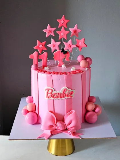 Come on Barbie, let's go party! This vibrant pink Barbie-themed cake features striped buttercream, a big fondant bow, and the classic Barbie logo, perfect for a stylish celebration.