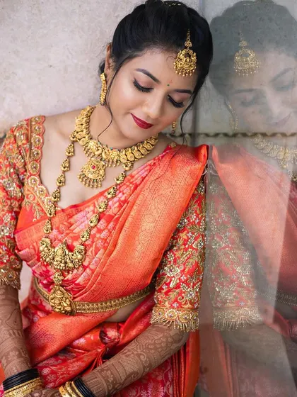 This is an artistic bridal portrait that captures the soft, glowing makeup beautifully. The elegant pose and the reflection highlight the flawless finish and the classic red lip, perfect for a South Indian wedding.