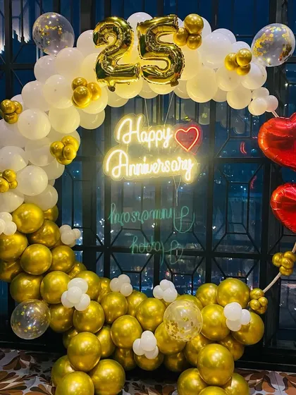 An elegant 25th-anniversary decoration with a balloon ring in gold and white, red heart foil balloons, and a "Happy Anniversary" neon sign.
