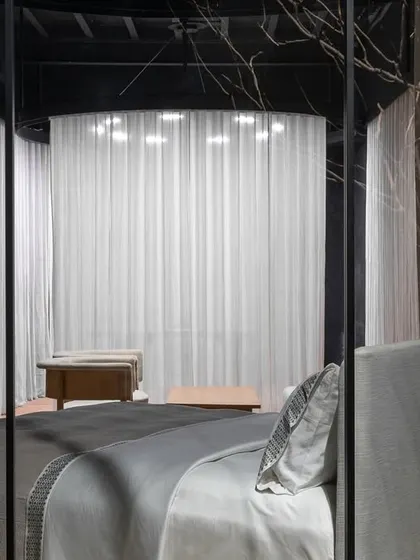 This view frames the bed display as a serene composition. Seen through the store's structural elements, the bed appears as a sanctuary, reinforcing the brand's ethos of quiet luxury and the home as a place of retreat.