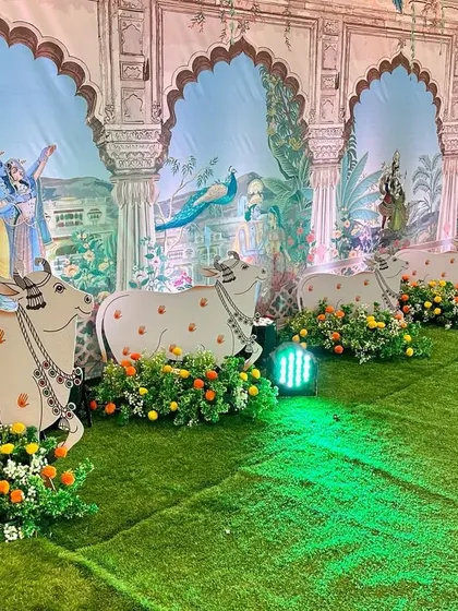 Another angle of the Pichwai-themed decor, focusing on the detailed artwork on the wall panels. The scene depicts Krishna and peacocks in a lush landscape, enhanced by the foreground arrangement of cow cutouts and marigolds.