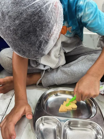 A child carefully decorates his freshly baked cookie with green icing, focusing intently on his delicious creation.