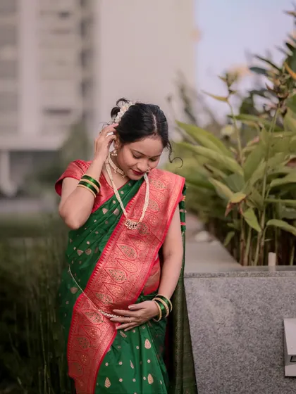 A graceful portrait of an expecting mother in a traditional green and red silk saree. This outdoor maternity photo captures her adjusting her hair, showcasing the intricate details of her attire and the beauty of pregnancy.