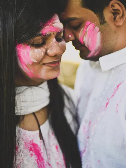 An intimate pre-wedding or post-Holi photoshoot moment. The couple is close together, their faces smeared with pink color, creating a playful and romantic image.