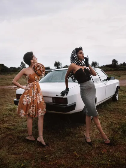 A wide shot from the 'BLANCH' series, establishing the scene. The two models, styled in distinct but complementary retro outfits, interact with the vintage car to build a complete visual story.