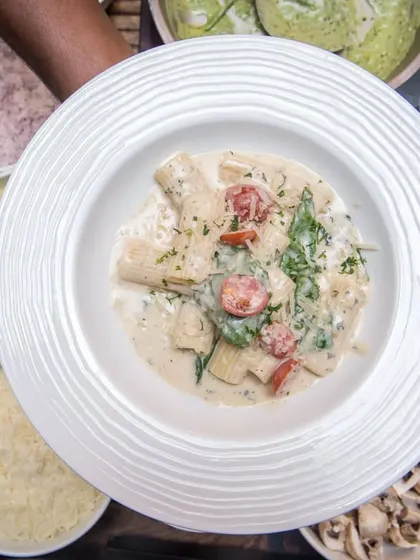 A top-down view of a creamy pasta dish surrounded by bowls of fresh ingredients like cherry tomatoes, spinach, and mushrooms.