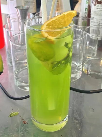 A bright green cocktail garnished with fresh mint and an orange slice. The color contrast is visually stunning.