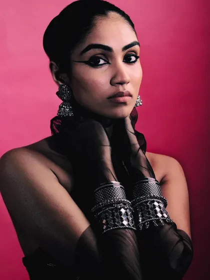 A direct and confident portrait from the 'Silver Jewels of Bombay' series. The model's bold eyeliner and traditional silver jewelry create a powerful statement against the pink background.