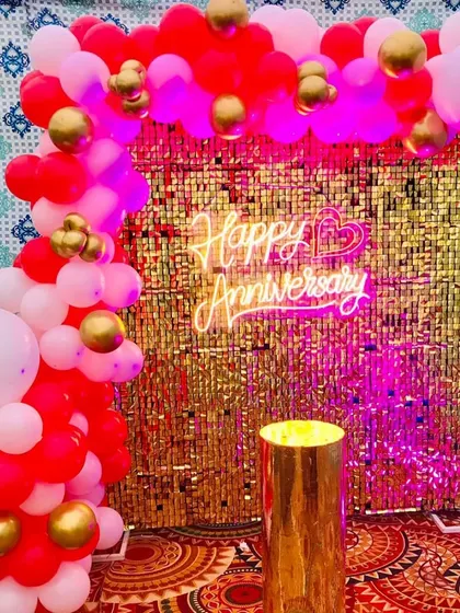 An anniversary decoration with a gold shimmer curtain backdrop. The setup includes a red, white, and gold balloon arch and a "Happy Anniversary" neon sign.