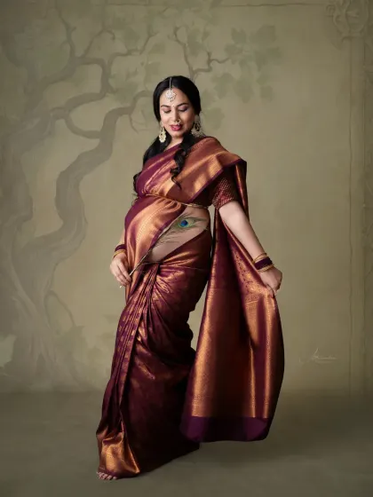A serene and artistic portrait. The mom-to-be, in her shimmering maroon saree, is pictured with a peacock feather, a symbol of grace and beauty.