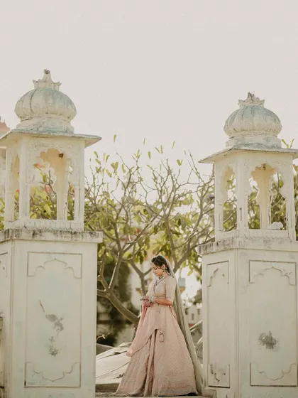 A portrait that feels like a painting. The bride stands between two traditional structures, her pastel lehenga complementing the soft light and serene architecture.