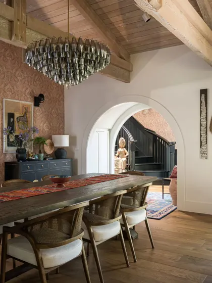 The dining room in the Saratoga home, viewed from the side. The image shows the layering of elements, from the patterned wallpaper and the large chandelier to the rustic dining table and the 'Devi' sculpture visible in the background.