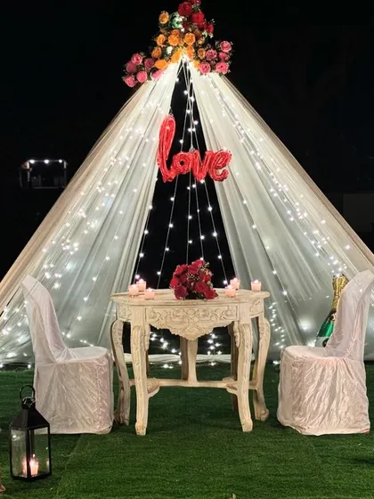 An intimate and romantic proposal setup in a garden, featuring a beautiful canopy tent draped in fairy lights, a 'Love' balloon, and a candlelit table for two.