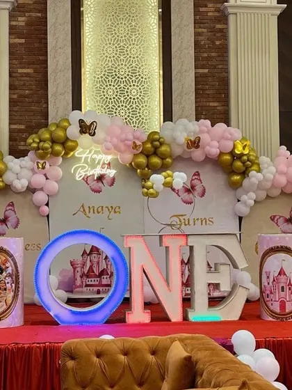 A lavish princess-themed first birthday party for Anaya. The stage decoration includes multiple backdrops, balloon arrangements, and large light-up "ONE" letters.