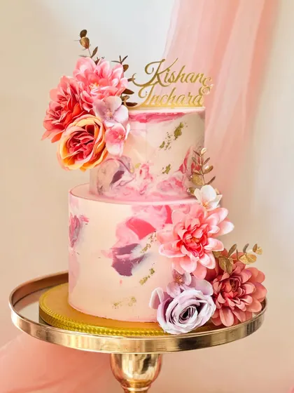 A two-tier cake with a beautiful pink and purple marble buttercream effect, flecked with gold leaf. It's decorated with an arrangement of silk flowers and a custom gold acrylic name topper.