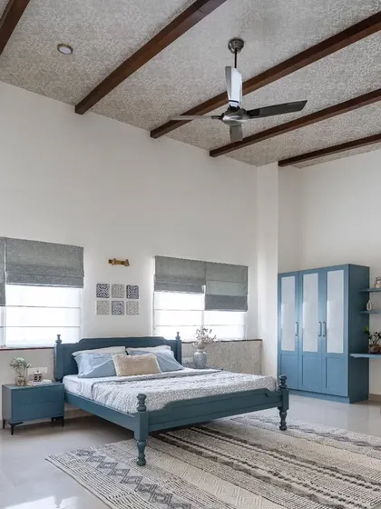 The master bedroom in the 'Spanish Country Home' features a serene blue color palette and a sloped roof with exposed rafters. Despite its spaciousness, the room feels cozy and intimate, complete with a custom study nook and wardrobe.