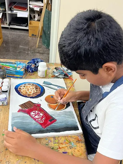 A young artist works on his "Food on the Table" assessment piece, carefully painting a KitKat bar and other treats. This project tests his understanding of light, shadow, and realism.