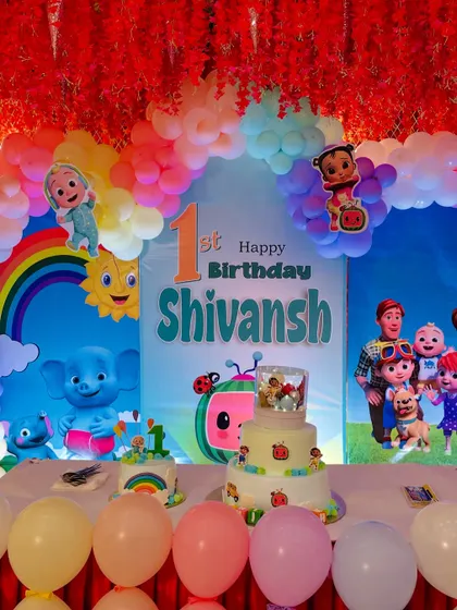 A close-up of a Cocomelon themed first birthday stage. The backdrop is filled with characters from the show, and the stage is decorated with a multi-layered cake and a rainbow balloon arch.